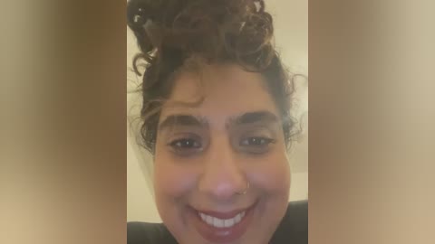 Media: Video of a smiling young woman with medium brown skin, curly brown hair, and dark eyebrows. She wears a black top, and the background is a blurred beige wall.