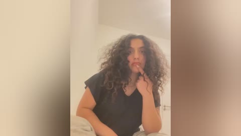 Media: Video of a woman with curly black hair, wearing a black t-shirt, sitting on a bed, biting her finger, blurred background.