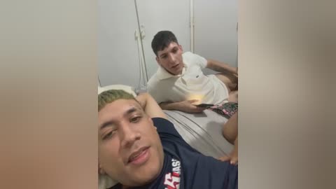 Media: A video of two young men, one with short blonde hair, lying on a bed in a dimly lit room, wearing dark shirts and shorts, with a mirror in the background.