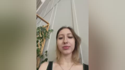 Media: Video of a young Caucasian woman with light skin, straight brown hair, and a neutral expression, wearing a black top. She stands in a minimalist room with a hanging plant and white walls.