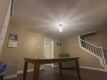 Media: Video of a bare, beige-walled, unfinished basement with a wooden table, white door, white railing staircase, and a sunken ceiling light.