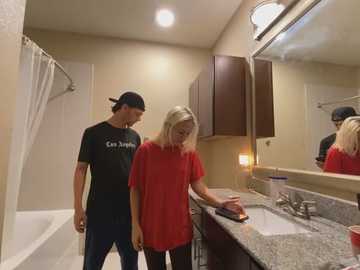 Media: Video of a blonde woman in a red shirt washing dishes in a beige bathroom with a man in a black shirt and cap standing nearby.