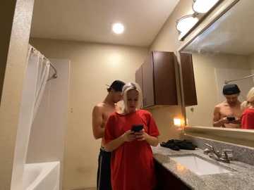 Media: Video of a bathroom with a shirtless man in dark pants, holding a phone, behind a woman in a red shirt, both focused on their phones; beige walls, granite countertop, mirror, and bathtub visible.