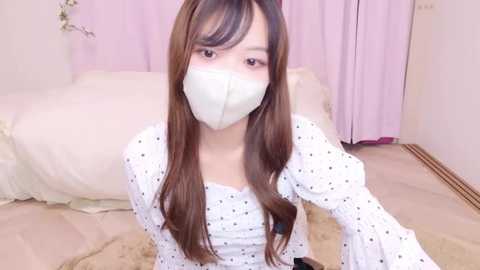 Media: Video of an Asian woman with long brown hair, wearing a white mask, a white polka-dotted dress, and a white shawl, kneeling on a beige carpet in a pastel room.