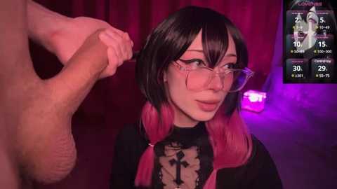 Media: A young Asian woman with short black hair and pink highlights, wearing glasses and a black and pink gothic-style top, is holding a man's hand. Background: red curtains, purple lighting, and a screen displaying a virtual reality game.