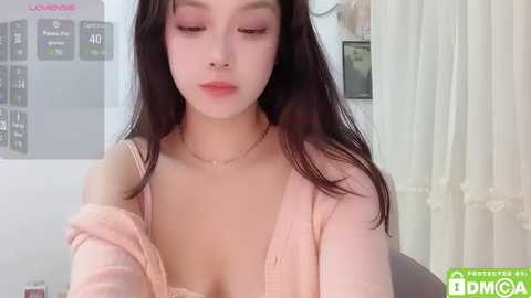 Media: Video of a young Asian woman with long black hair, wearing a pink cardigan, resting her head on her hand, in a bedroom setting with a weight scale and white curtains.