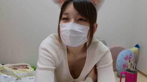 Media: Video of an Asian woman with light skin and straight black hair, wearing a white face mask, animal ear headband, and a beige shirt, leaning on a table. Background includes a blue container, white fabric, and a pink mug.