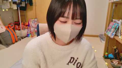 Video of an East Asian woman with straight black hair, wearing a white mask, light sweater, and a gray sweatshirt with \"Pio\" in black. Background shows a cluttered room with toys, books, and a wooden shelf.