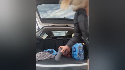 Media: Video of a blonde woman in a black jacket, bending over to retrieve a blue water bottle from the open trunk of a car.