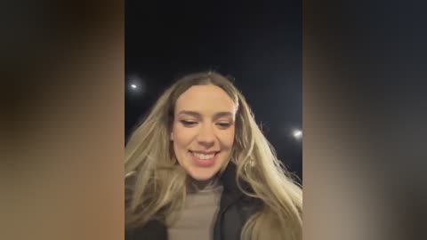 Media: Video of a smiling woman with long blonde hair, wearing a black jacket and light-colored top, set against a dimly lit, blurred background.