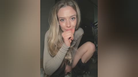 Media: Video of a young, light-skinned woman with long blonde hair, wearing a beige long-sleeve top, black shorts, and black socks, crouching in a dimly lit room, with a blurred background.