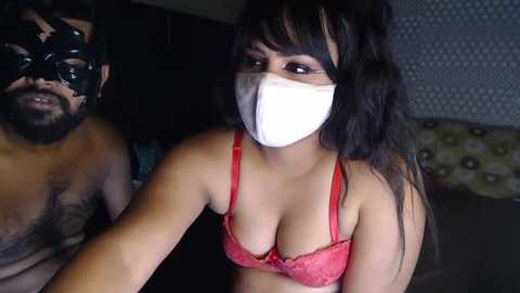 Media: Video of a dark-skinned man in a black mask, and a light-skinned woman with long dark hair in a red lace bra, both wearing white face masks, indoors.