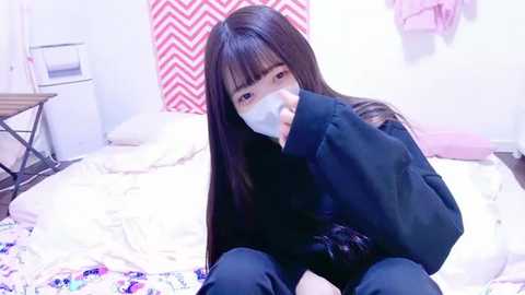 Media: A video of an East Asian woman with long black hair, wearing a surgical mask and dark clothing, sitting on a bed with white bedding, in a room with pink and white chevron-patterned curtains and a wooden chair.