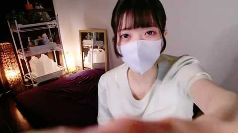 Media: Video of a young Asian woman with a light complexion, wearing a white surgical mask, sitting on a bed with a dark purple blanket, in a dimly lit room with a bookshelf and wicker basket in the background.