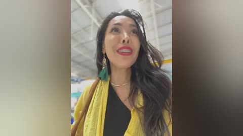 Media: Video of a smiling Asian woman with long black hair, wearing a mustard-yellow cardigan over a black shirt, standing in a brightly lit, industrial-looking room with white ceilings and pipes.