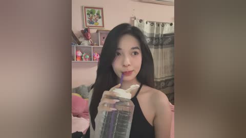 Media: Video of an Asian woman with long black hair, wearing a black sleeveless top, drinking from a clear plastic bottle in a pink bedroom with toys, a framed picture, and a curtain.