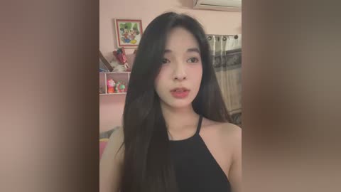 Media: A video of an East Asian woman with long black hair, wearing a black halter top, standing indoors in a softly lit room with pink walls, shelves, and curtains.