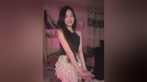 Media: A video of an Asian woman with long black hair, wearing a black sleeveless top and a white skirt with floral patterns, standing in a cluttered room with pink lighting.