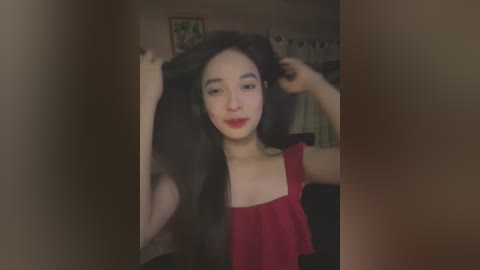 Media: Video of an Asian woman with long, straight black hair, wearing a red top, holding her hair. Background shows a dimly lit room with a framed picture and white curtains.