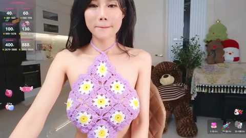 Media: Video of an East Asian woman with long black hair, wearing a purple crocheted halter top adorned with white and yellow daisies. Background includes a plush teddy bear and a plush toy.