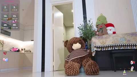 Media: Video of a plush teddy bear in a cozy, modern living room with a white door open, showcasing a kitchen with pink accents and a calendar displaying December.