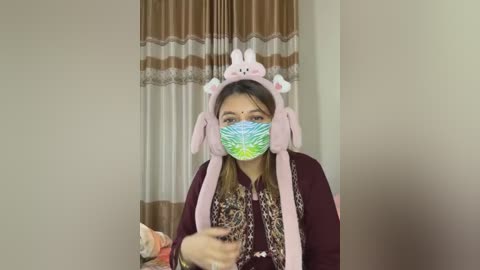 Media: Video of a young woman in a pink bunny mask, green face mask, and maroon sweater, holding a red gift bag, indoors with beige walls and brown curtains.