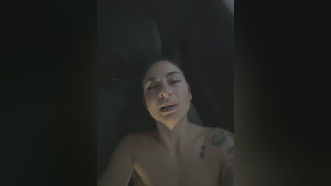 Media: Video of a nude woman with fair skin and long black hair, lying on her back in a dark, dimly lit room. She has tattoos on her shoulder and is visibly distressed, eyes closed, mouth open.