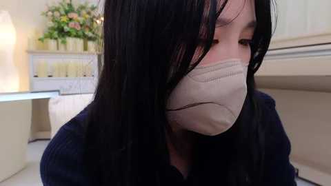 Media: A video of an East Asian woman with long black hair, wearing a beige surgical mask, sitting indoors, with a floral arrangement and a white dresser in the background.