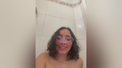 Media: Video of a young, smiling woman with shoulder-length wavy black hair, wearing clear, round glasses, standing in a tiled shower with white walls and a decorative border. She is topless.