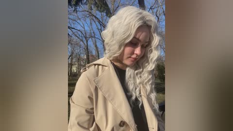 Media: Video of a young woman with wavy platinum blonde hair, wearing a beige trench coat, standing outdoors in a park with bare trees and a clear blue sky.