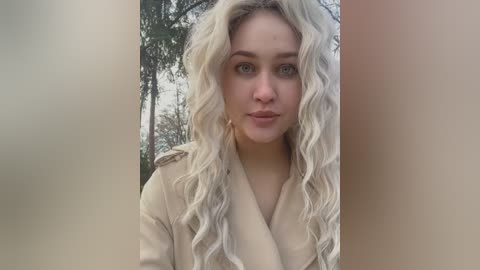 Media: Video of a fair-skinned woman with long, wavy platinum blonde hair, wearing a beige coat, standing in front of a blurred forest background.