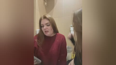 Media: Video of a woman with long, straight, light brown hair, wearing a red sweater, looking in a bathroom mirror, with another person partially visible.