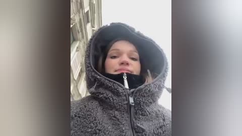 Media: A video of a young girl in a gray, fuzzy hooded jacket, her face partially obscured by the hood. She is standing in front of a wooden door with a white background.