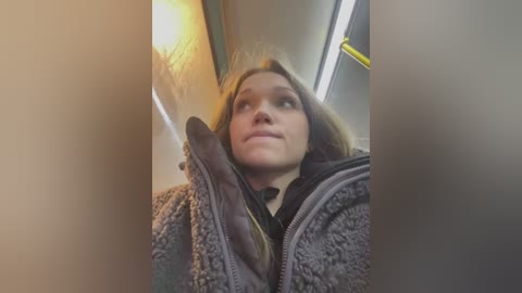 Video of a young woman with light skin and straight, shoulder-length blonde hair, wearing a grey, textured jacket, looking up, blurred background of a subway car.