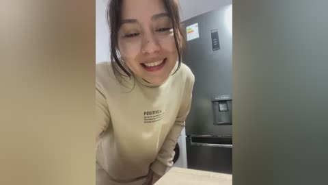 Media: Video of a smiling Asian woman with brown hair, wearing a beige sweater, bending over a counter in a modern, grey kitchen with a refrigerator.