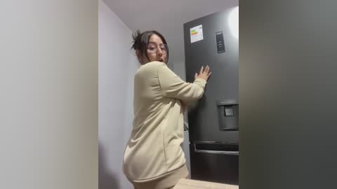 Media: Video of a woman in beige pajamas reaching into a dark metallic refrigerator in a small, plain room with white walls and wooden floor.