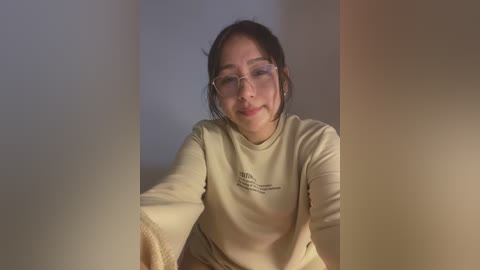 Media: Video of an East Asian woman with glasses, fair skin, and dark hair, wearing a beige sweatshirt with a small logo. She is smiling, sitting against a plain, beige wall, with soft lighting creating a warm, cozy atmosphere.