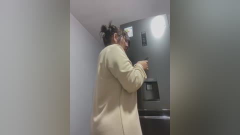 Media: Video of a woman with dark hair in a bun, wearing a beige coat, using a black ATM in a dimly lit, narrow corridor with white walls.