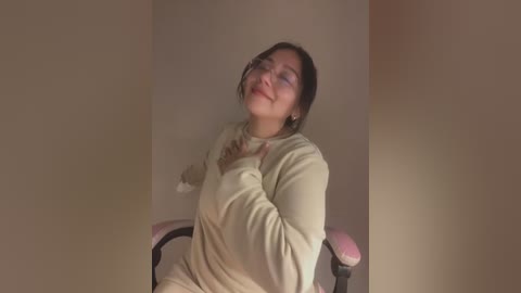 Media: Video of a smiling Asian woman with glasses and medium-length dark hair, wearing a beige sweater, sitting on a pink gaming chair against a plain beige wall.