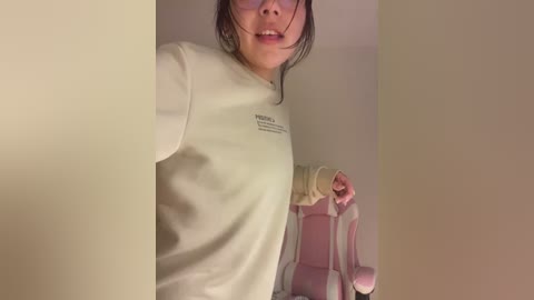 Media: A video of a young Asian woman with glasses, wearing a white long-sleeve shirt and pink headphones, taking a selfie in a dimly lit room.