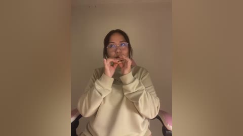 Media: A video of a person with glasses, medium build, light beige sweater, and brown hair, holding a small green object, in a dimly lit room with beige walls.