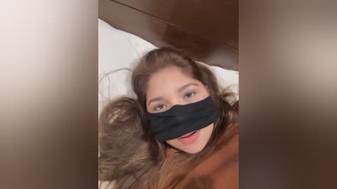 Video of a woman with long brown hair, wearing a black face mask and a brown sweater, lying on a white pillow against a wooden headboard.