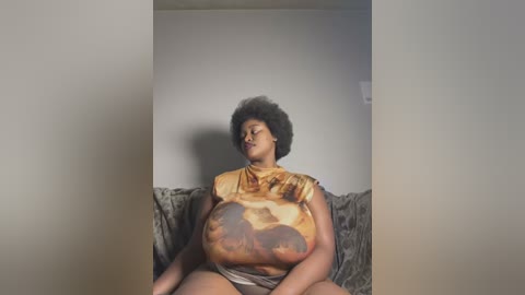 Media: A video of a pregnant Black woman with a voluminous afro, wearing a sleeveless, mustard-yellow top with a brown pattern, sitting on a gray couch against a plain white wall.