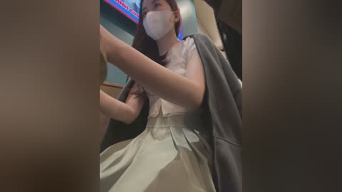 Media: A video of an Asian woman in a white dress and gray jacket, wearing a face mask, seated in a dimly lit room with a TV screen displaying red and blue colors in the background.