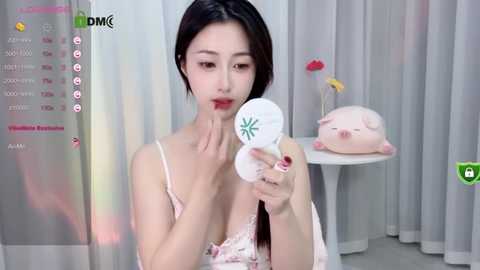 Media: Video of a young Asian woman with fair skin, black hair, and light pink lingerie, applying makeup in a minimalist, modern bathroom with white tiles and a pink piggy bank.