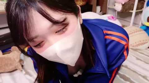 Video of an East Asian woman with long black hair, wearing a blue jacket, white mask, and glasses, lying on a bed with stuffed toys, in a cozy, cluttered room.