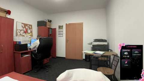 Media: Video of a small, sparsely furnished office with a computer desk, chair, wooden door, cork board, and a large red filing cabinet.