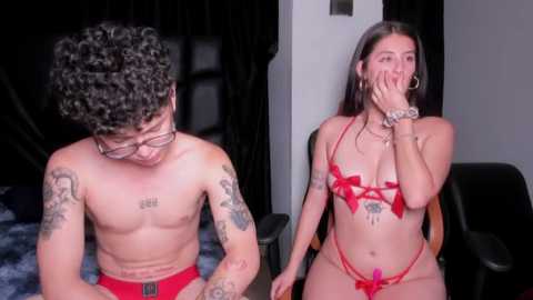 Media: Video of two young adults in a dimly-lit bedroom: a shirtless man with tattoos, wearing red briefs, and a woman in red lingerie, covering her face with her hand.