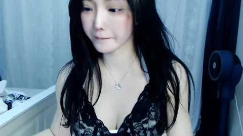 Media: Video of an East Asian woman with long black hair, fair skin, and a slender figure, wearing a black lace bralette, sitting in a dimly lit bathroom with a white bathtub and a blue speaker in the background.