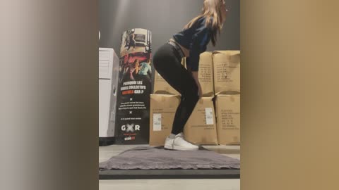 Media: A video of a woman with long hair, wearing a black hoodie, black leggings, and white sneakers, squatting on a gray mat in a gym with stacked cardboard boxes in the background.